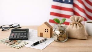 Wooden house model, jar with coins and plant, calculator, money, pen, sack, glasses, coffee cup, US flag background.