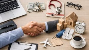 Two people shaking hands over a desk with laptop, money, keys, stethoscope, house model, clock, and coffee cup.